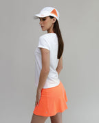 Woman wearing a white t-shirt, orange skirt, and white cap with orange logo on a gray background