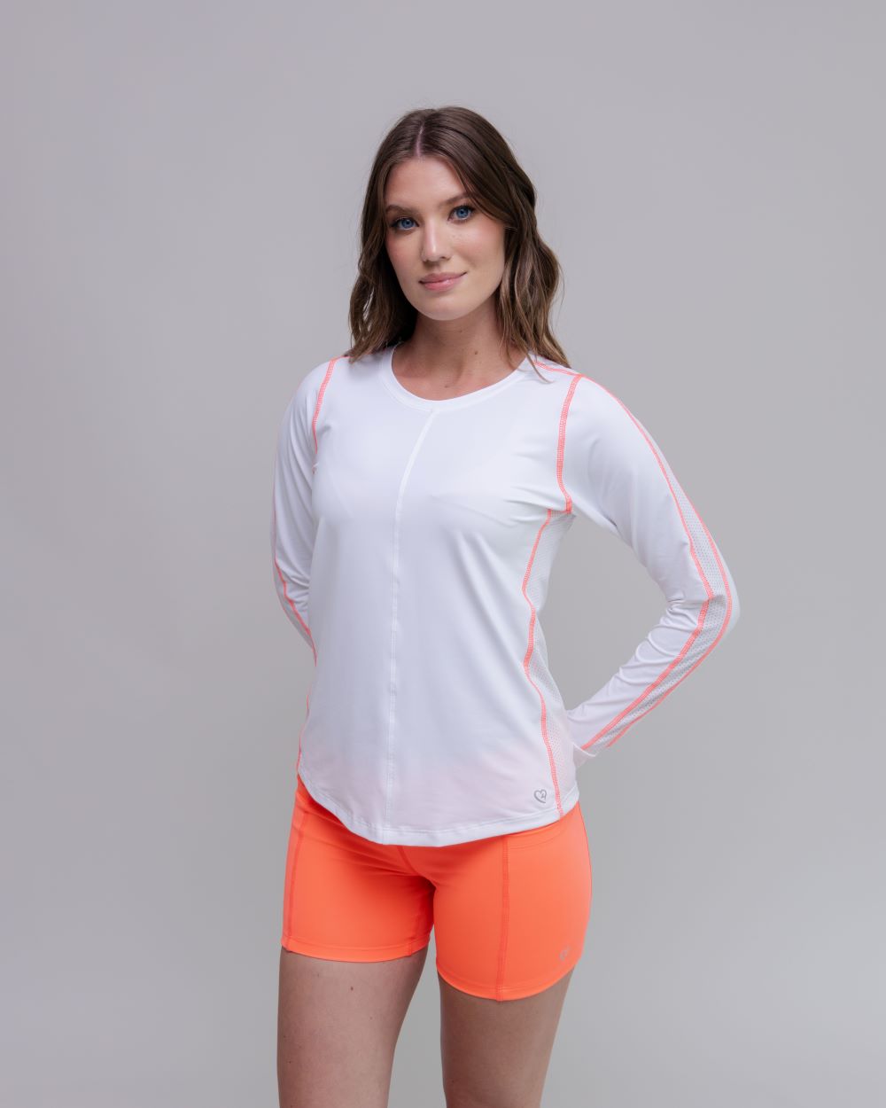 Woman wearing a white long-sleeve athletic top with pink accents and orange shorts on a gray background